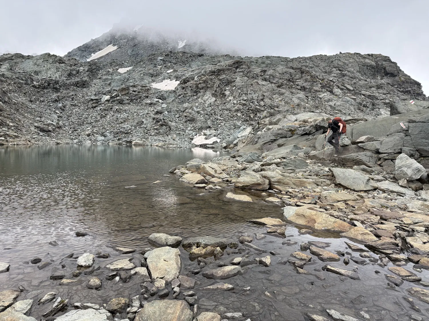 Man walking down rocks near lake with clouds close above